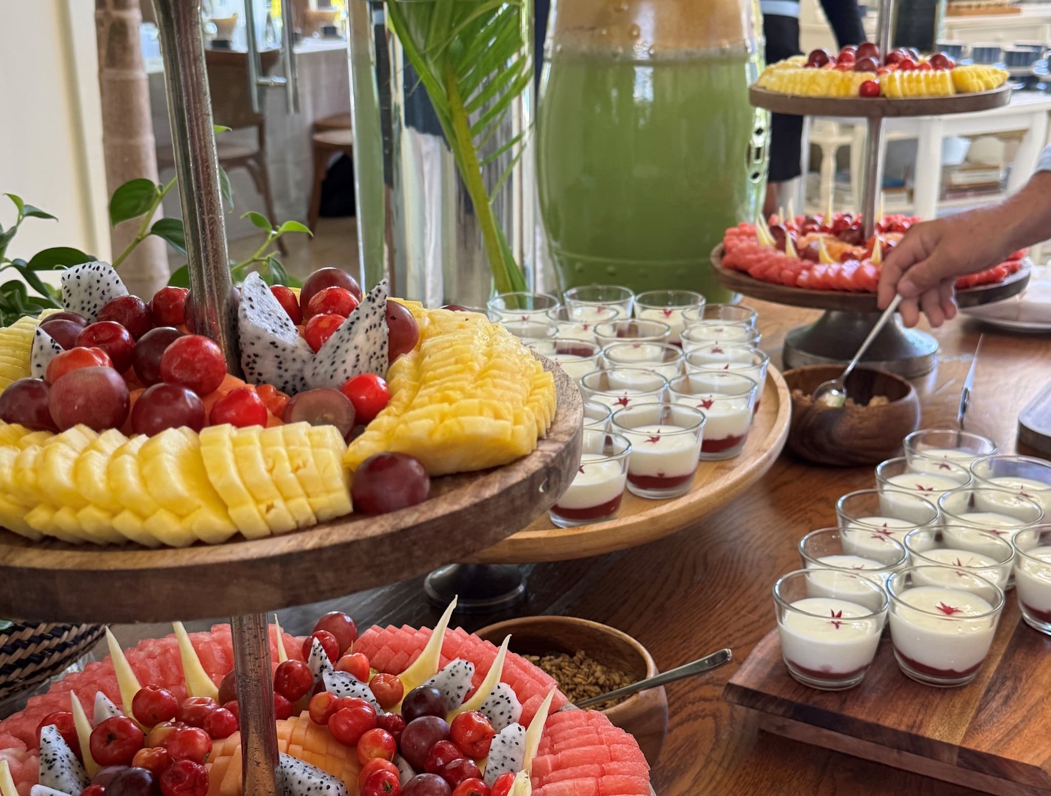 A vibrant buffet display featuring tiered wooden stands filled with fresh fruit, including pineapple, dragon fruit, grapes, watermelon, and cherries. Nearby, rows of small glass cups hold layered desserts topped with a delicate garnish. A large glass dispenser of green juice stands behind, while a hand reaches in to scoop toppings from a small bowl. The setting feels bright and fresh, with natural light and a polished wooden table enhancing the colorful presentation.