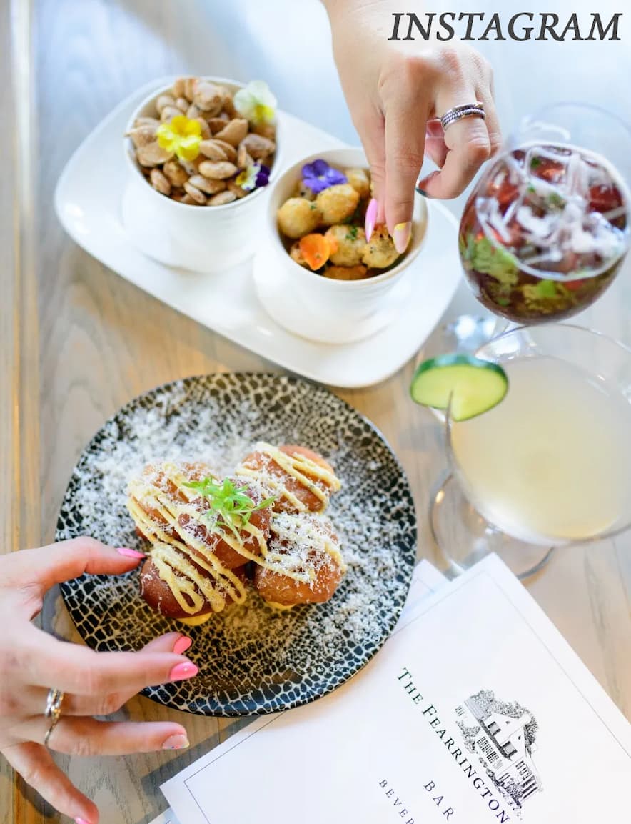 Overhead view of hands reaching for small plated bites and snacks at a restaurant table, with cocktails, a patterned plate of food, small bowls of appetizers, and a menu visible, creating a bright, social dining moment.