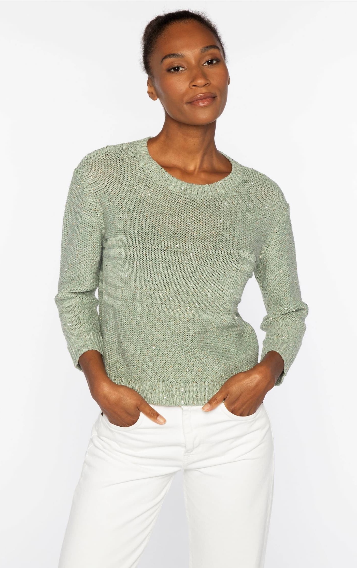 Woman wearing a sage-green knit sweater with subtle texture, paired with light trousers against a neutral background.