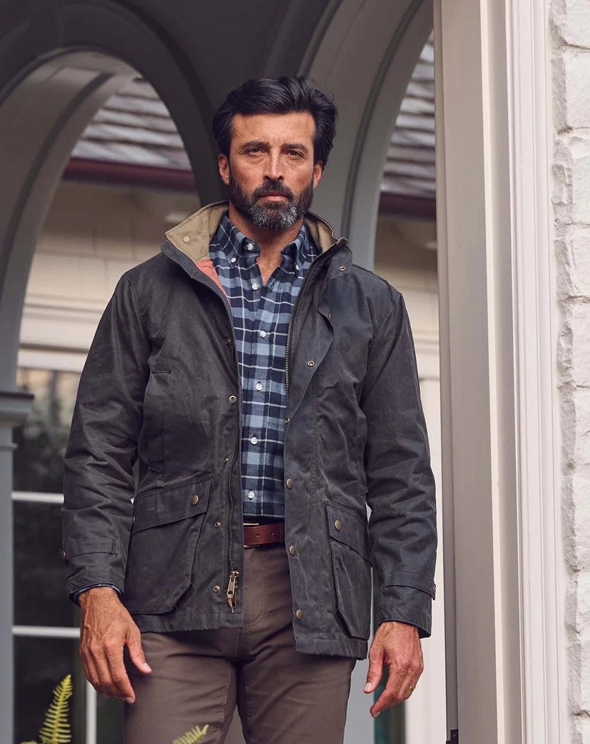 A man with dark hair and a beard stands in a doorway, wearing a black jacket over a plaid shirt and brown pants. The background features white brick walls and an arched window. Fearrington Village