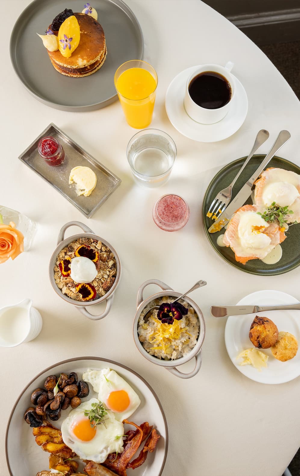 Overhead view of an elegant brunch spread featuring smoked salmon eggs Benedict, pancakes with fruit and cream, granola and oatmeal bowls topped with edible flowers, bacon and eggs, pastries, fresh juice, coffee, and colorful breakfast cocktails arranged on a white tablecloth.