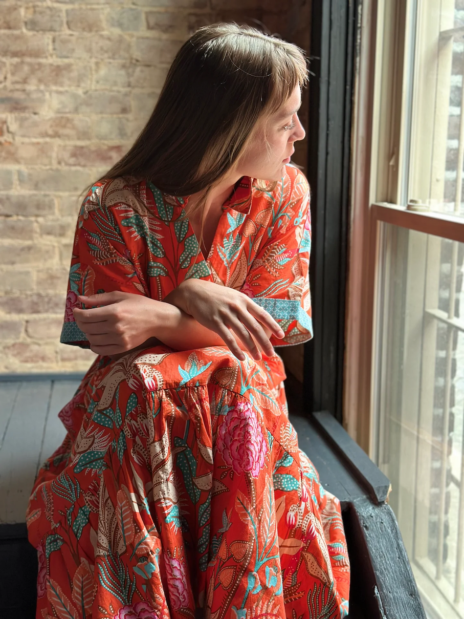 Woman in a vibrant red patterned dress sitting by a window, looking outside with soft natural light and a brick wall in the background.