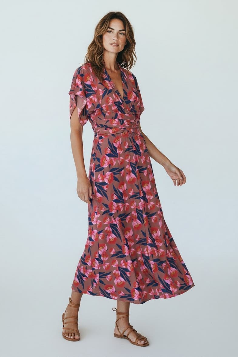 A woman wears a flowing wrap-style dress in a warm rose tone with vibrant floral prints in pink and deep blue. The dress features soft flutter sleeves, a cinched waist tie, and a graceful mid-length hem that moves easily. Paired with simple leather sandals, the look feels feminine, relaxed, and polished.