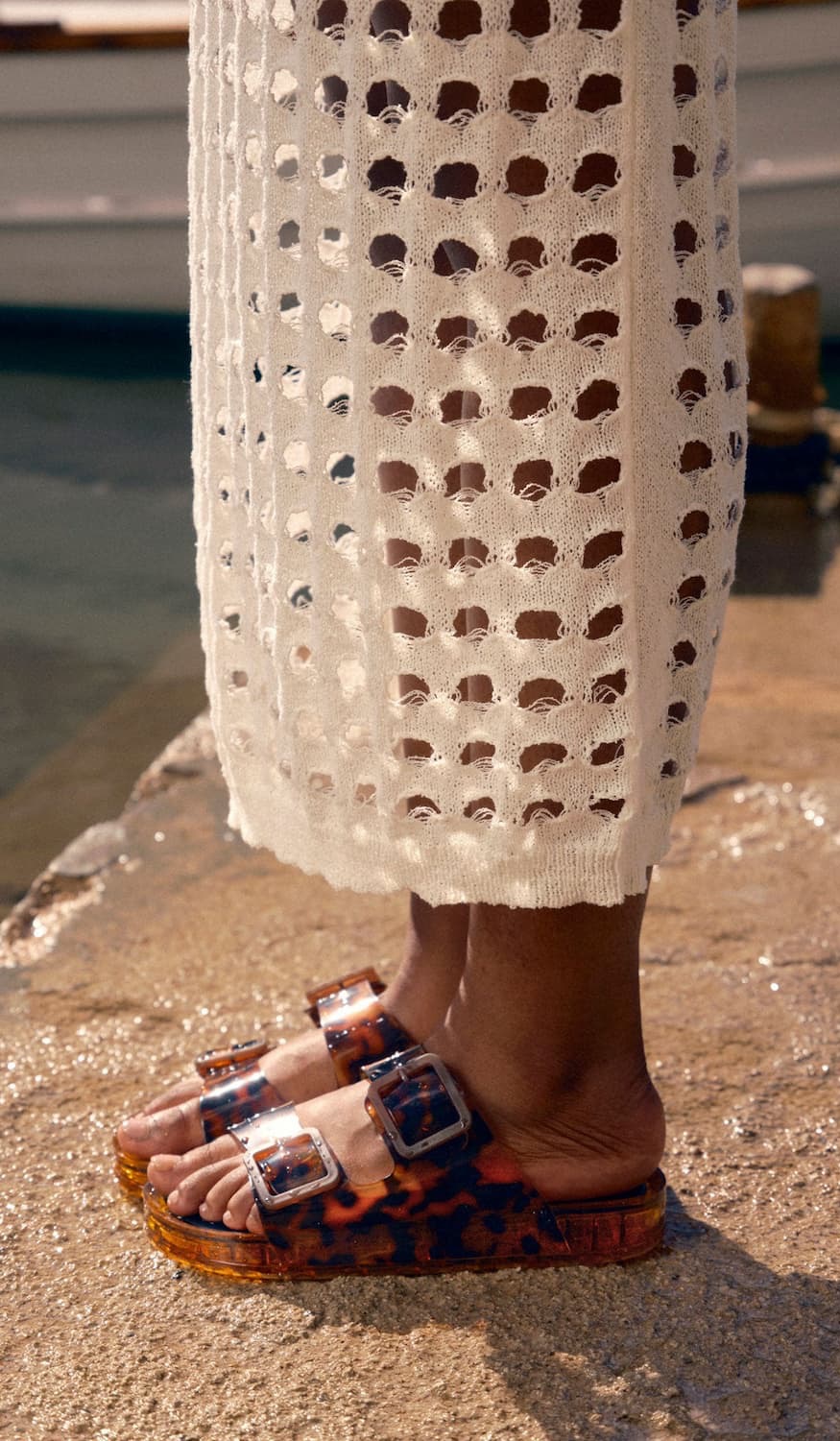 Close-up of a person standing poolside wearing a cream open-knit crochet cover-up that falls to the ankles and translucent tortoiseshell double-strap slide sandals with oversized silver buckles. Sunlight highlights the textured knit and glossy sandals, creating an effortless resort-inspired look.