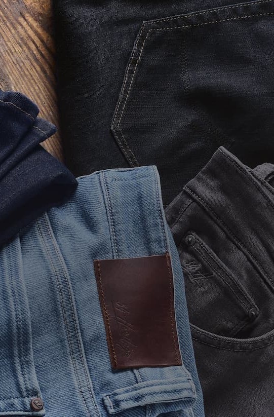 Several pairs of folded jeans in different shades of blue and black are arranged on a wooden surface. Visible details include pockets, stitching, and leather patches on the waistbands. Fearrington Village