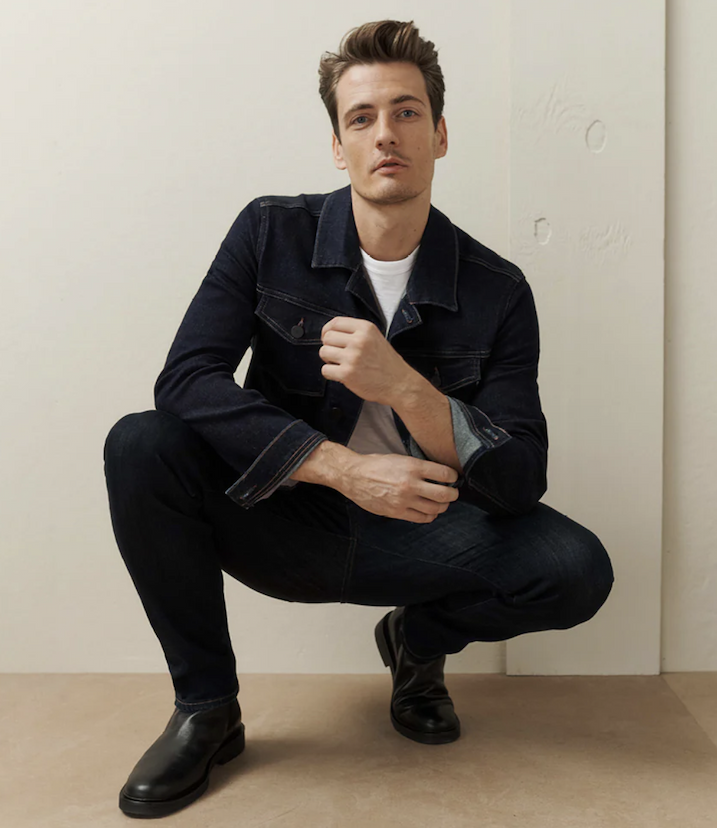 A man with short brown hair squats indoors, looking at the camera. He wears a dark denim jacket over a white T-shirt, dark jeans, and black boots. The background is simple and neutral. Fearrington Village