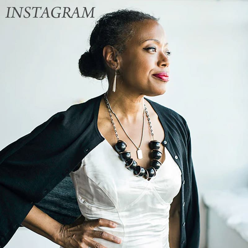 Woman in a white dress and black cardigan wearing a bold black beaded necklace and long earrings, standing in soft natural light and looking off to the side.