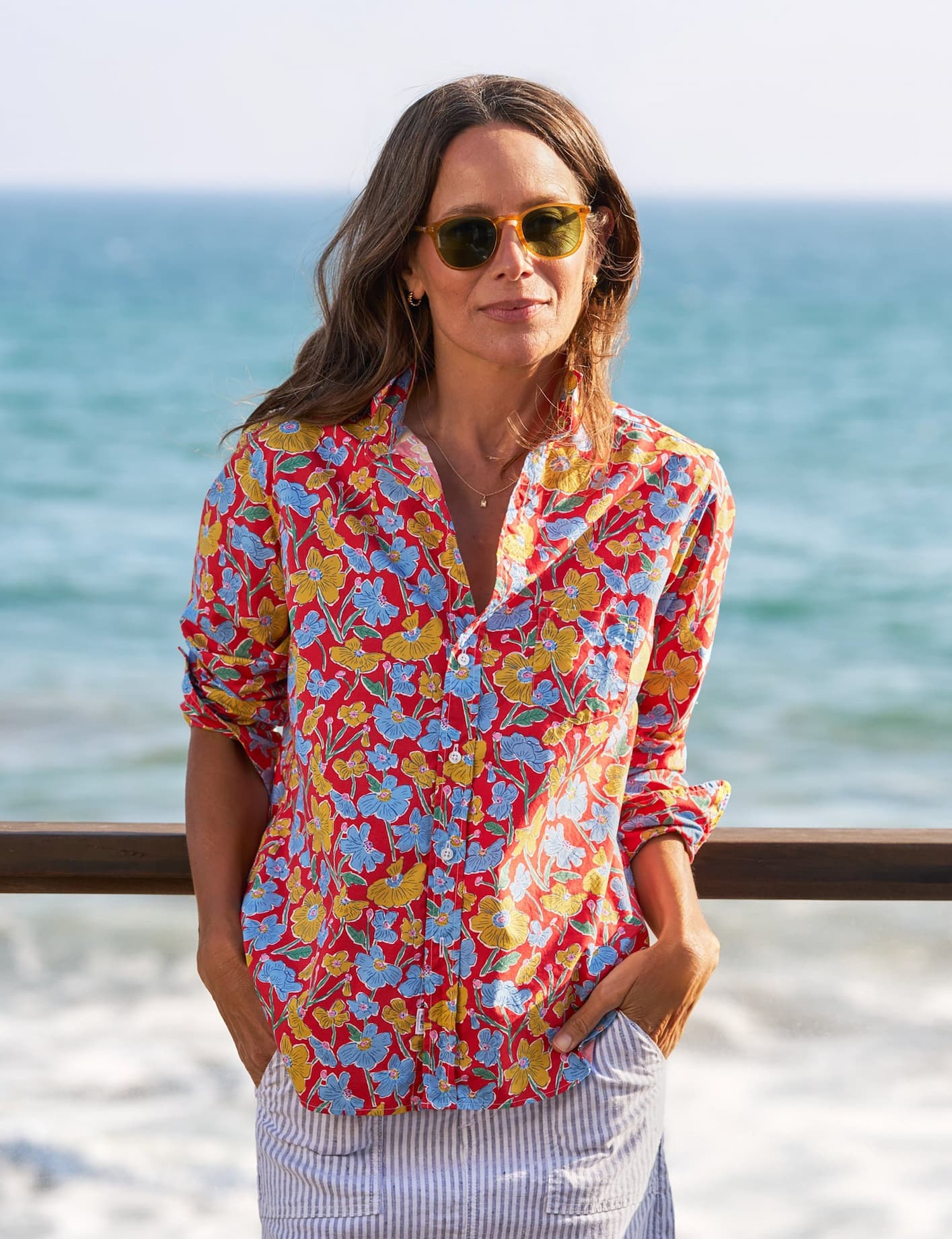Woman in a vibrant red floral blouse and striped skirt stands by a wooden railing overlooking the ocean, wearing sunglasses and looking toward the camera in warm coastal light.