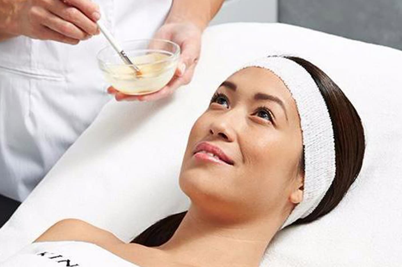 A woman relaxes during a facial treatment while a spa therapist prepares a skincare mixture in a small bowl beside the treatment bed.