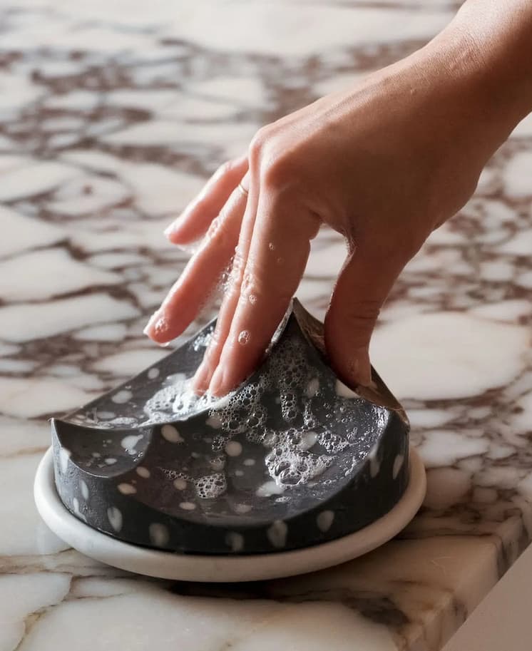 A hand lathers a black-and-white speckled Ume Erode soap on a small white dish, resting on a marble countertop.