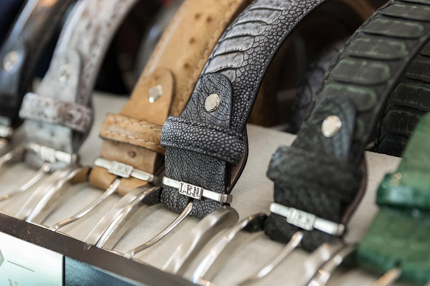 Display of handcrafted leather belts in varied textures and colors, each with a polished metal buckle, arranged on a shop counter.