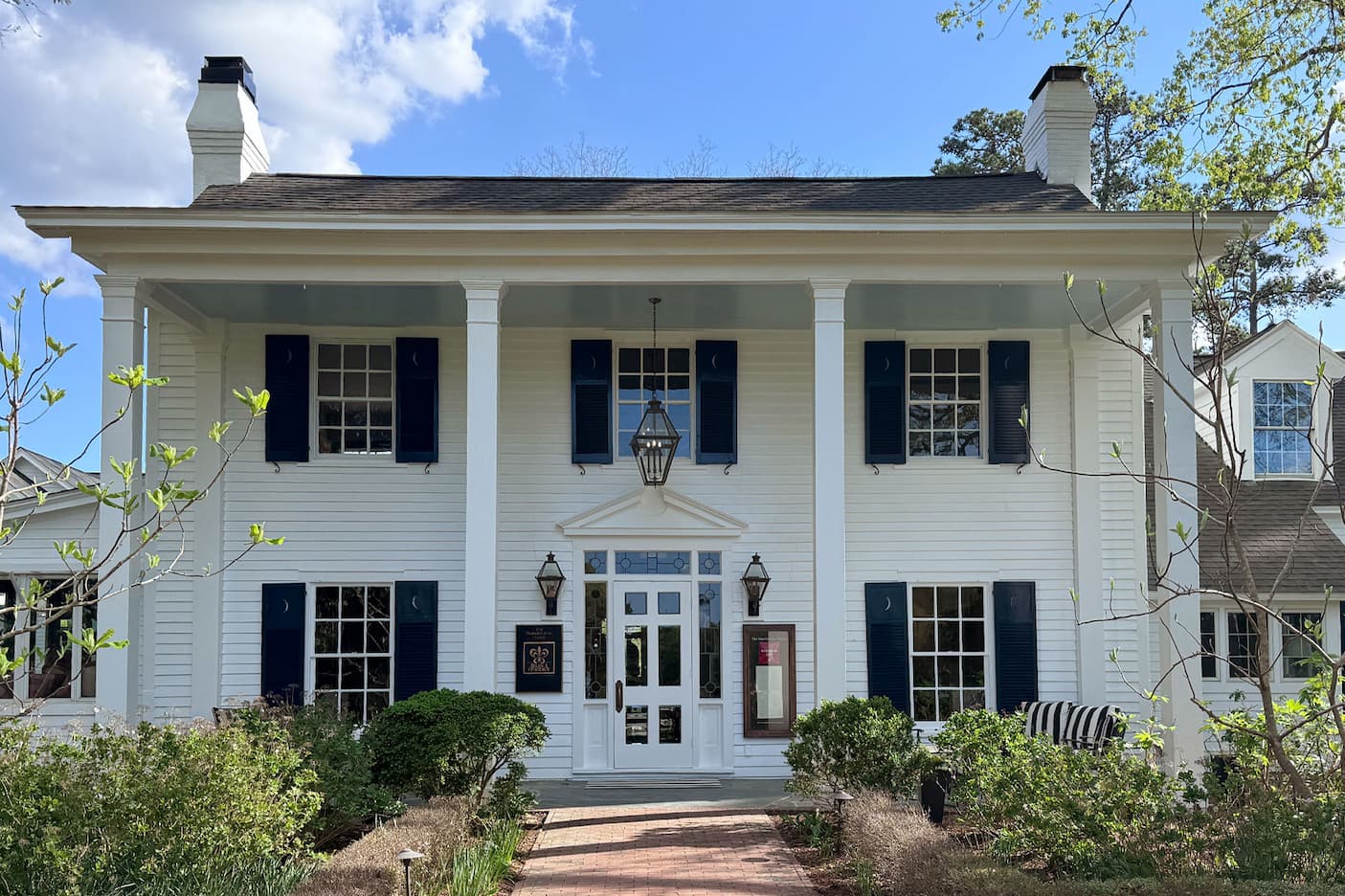 White, two-story colonial-style building with a wide front porch supported by columns, black shutters on symmetrical windows, and twin chimneys. A brick path leads to a central glass-paneled door flanked by lanterns, with neatly trimmed shrubs and early spring greenery under a blue sky.