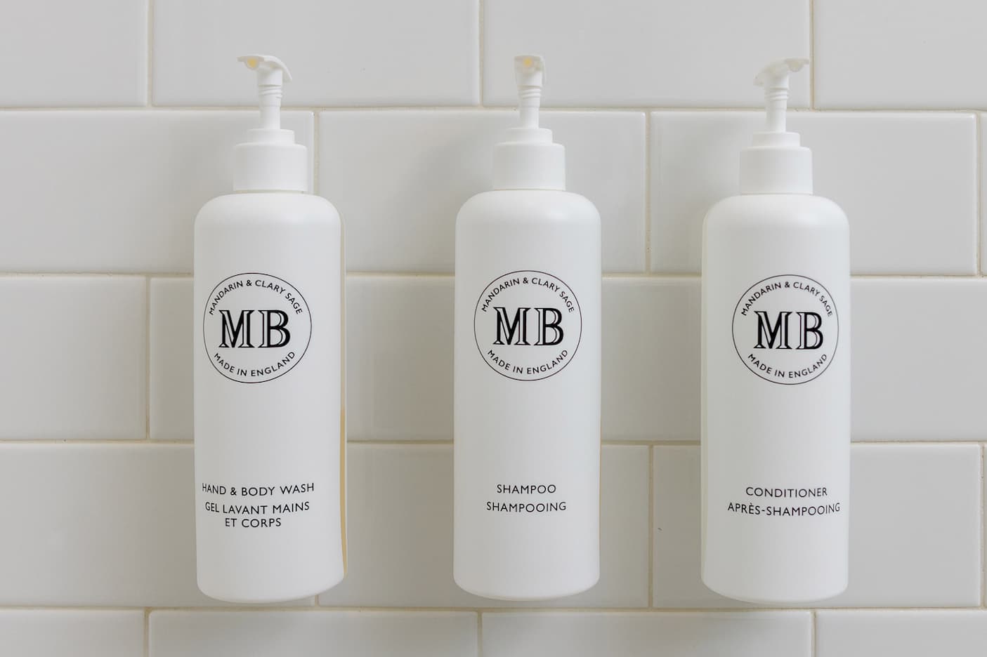Three white pump bottles labeled hand & body wash, shampoo, and conditioner arranged in a row against a clean white tiled wall, with minimalist branding.