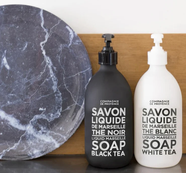 Two Marseille liquid soaps in matte black and white bottles labeled “Black Tea” and “White Tea,” styled beside a gray marble tray against a warm wood backdrop for a clean, modern look.