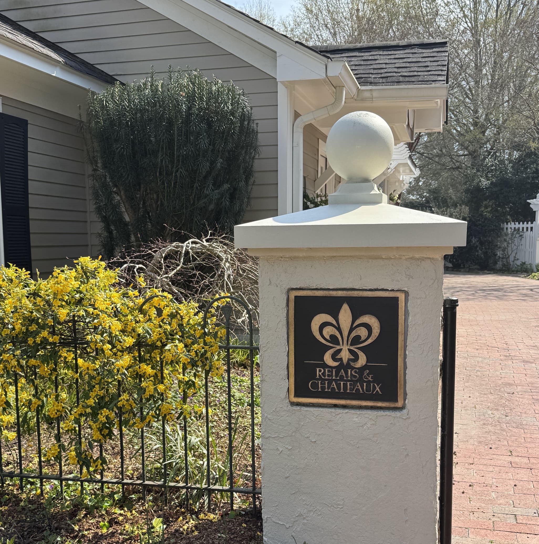 Garden entrance with a Relais & Châteaux plaque mounted on a white pillar, surrounded by spring plantings and a brick walkway leading toward the inn.