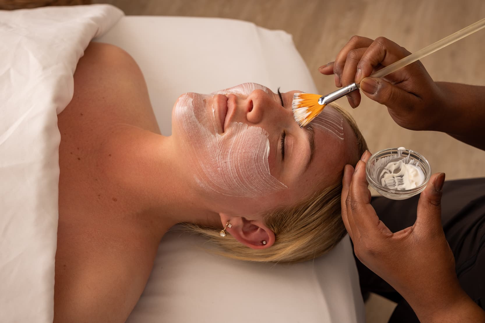 Person lying on a spa bed receiving a facial treatment, with a therapist applying a creamy mask to the face using a brush while holding a small bowl of product.