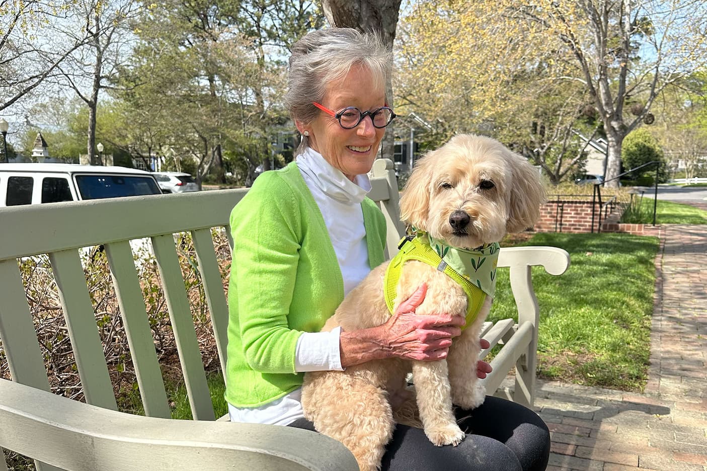 Woman in a green sweater sitting on a bench outdoors, smiling while holding a small fluffy dog wearing a green harness in a sunny neighborhood setting.