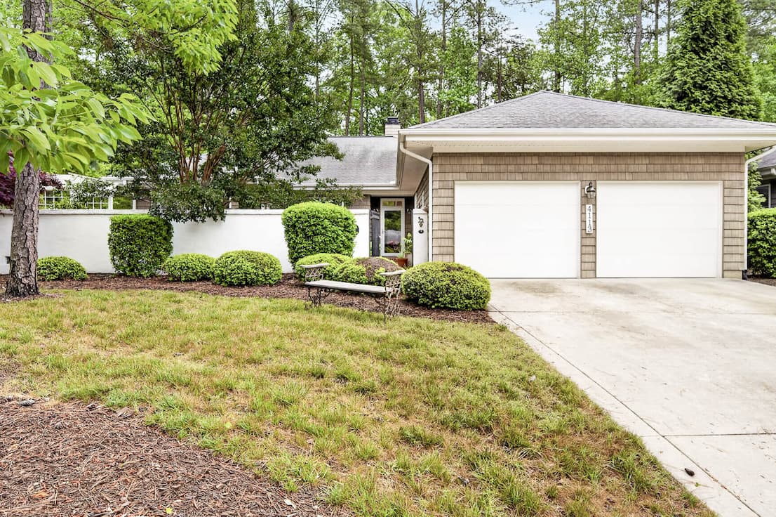 A single-story home with a two-car garage sits behind a driveway, surrounded by neatly trimmed shrubs, a small lawn, and mature trees in a quiet residential setting.