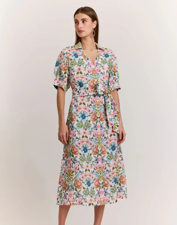A woman models a short-sleeve shirt dress in a soft floral print, styled with a tie waist and simple hoop earrings against a neutral background.