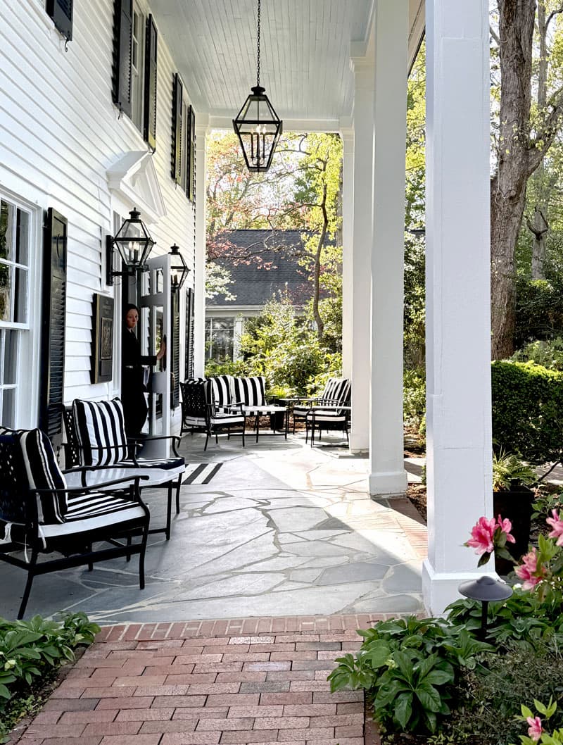 Shaded front porch with tall white columns, black shutters, and hanging lanterns. Black-and-white striped cushioned chairs line the stone floor, with a brick walkway leading up. A person opens the front door, and greenery and blooming flowers frame the scene.