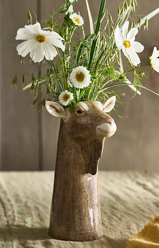 Ceramic goat-shaped vase holding white cosmos and small daisy-like flowers arranged with airy green stems on a linen-covered table.