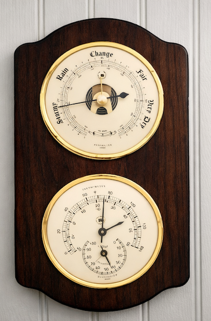 Wall-mounted wooden weather station with brass-trimmed analog dials, showing a barometer labeled Stormy to Fair and a lower dial measuring temperature and humidity.