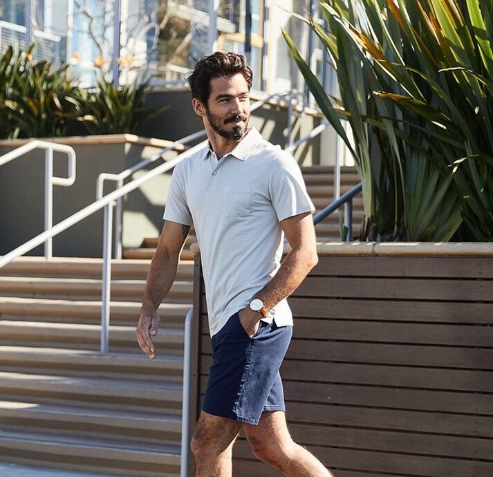 Man walking along a city sidewalk wearing a light polo, navy shorts, and sneakers, styled in a clean, casual summer look.