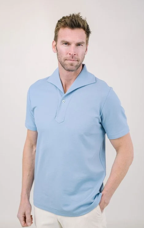 Model wearing a light blue short-sleeve knit polo with an open collar and white pants against a neutral background. Fearrington Village.