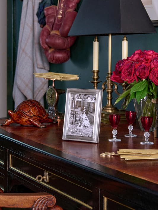 Silver photo frame displayed on an elegant console with red roses, brass candlesticks and ruby glassware. Fearrington Village.