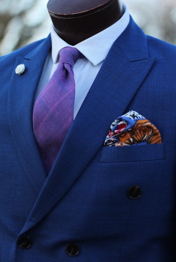 Close-up of a tailored blue double-breasted suit jacket styled with a white shirt, purple tie, patterned pocket square, and lapel pin on a mannequin.