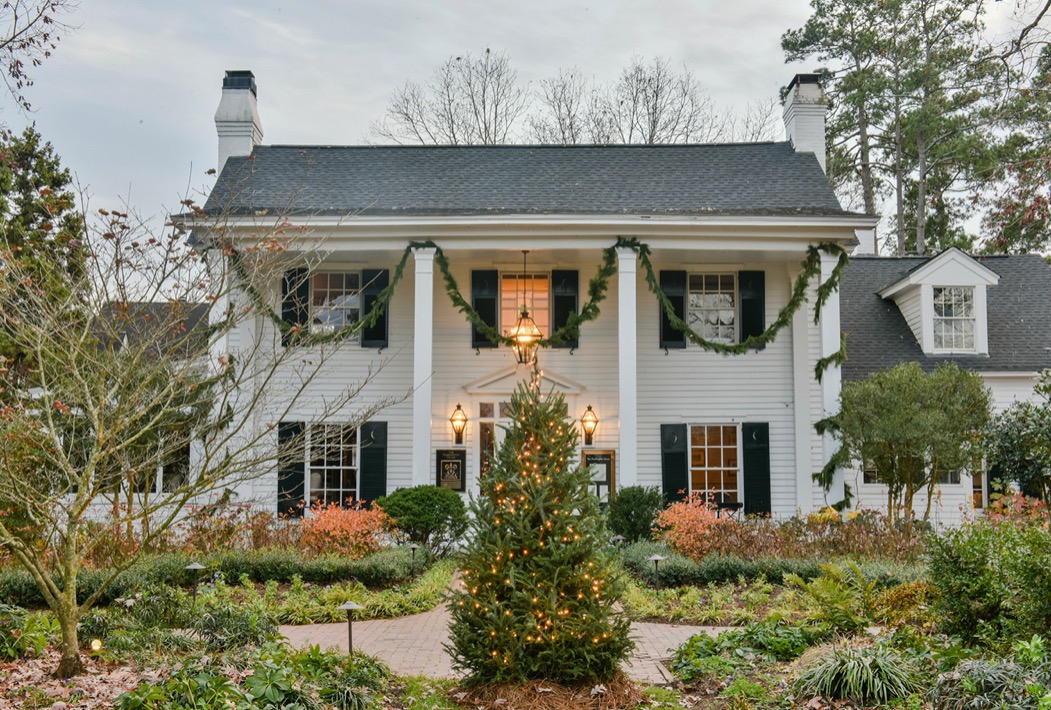 The Fearrington House Inn is shown from the front, decorated for the holidays with garland draped across the porch, glowing lanterns, and a lit Christmas tree centered along the garden path.
