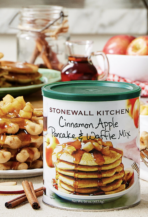 A canister of Stonewall Kitchen Cinnamon Apple Pancake & Waffle Mix is on a table with pancakes, diced apples, syrup, cinnamon sticks, and apples in the background. Fearrington Village