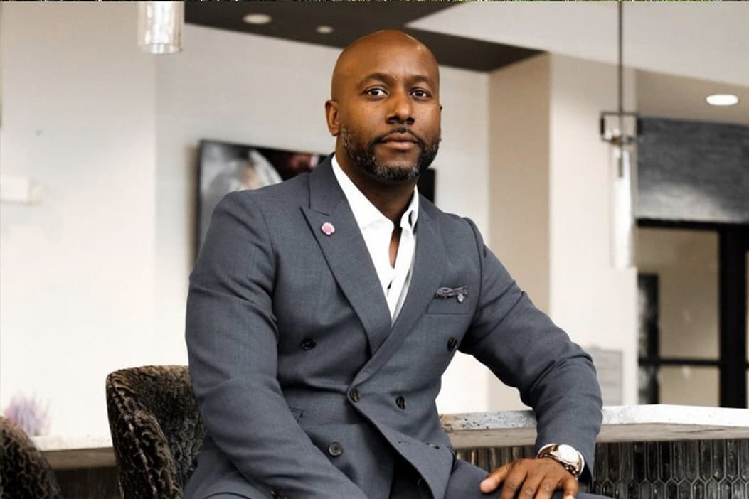 Well-dressed man seated indoors, wearing a tailored gray double-breasted suit with a white shirt and pocket square, looking confidently toward the camera in a modern interior setting.
