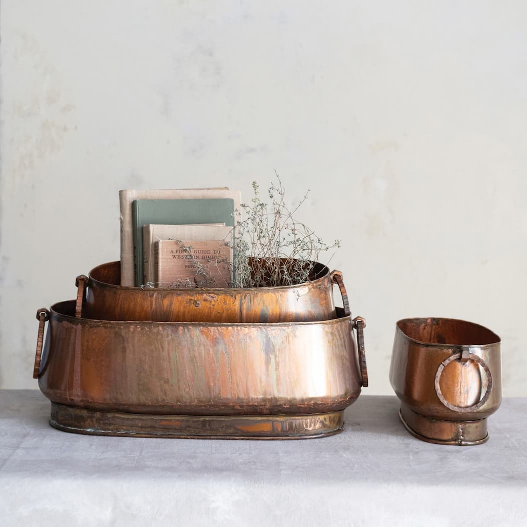 Set of weathered copper planters in varying sizes with side handles, styled on a neutral surface against a soft plaster wall, holding books and delicate dried stems for a warm, rustic decorative display.
