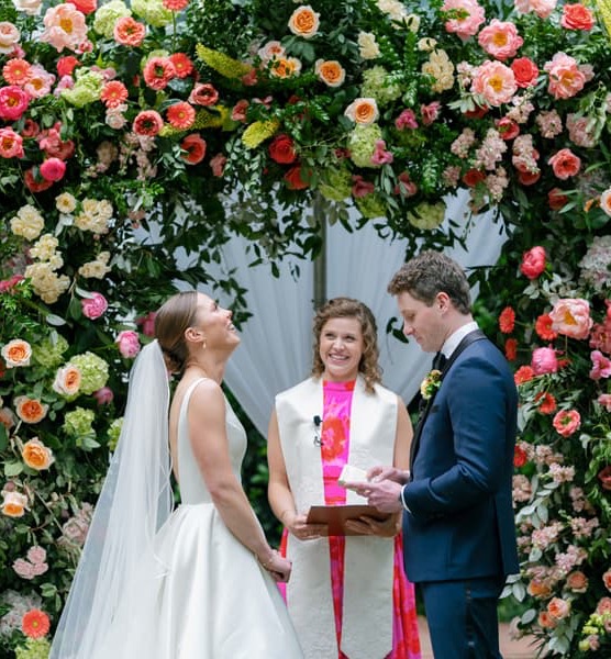 Bride and groom exchange vows beneath a lush floral arch filled with pink, coral, peach, and white blooms. Fearrington Village.