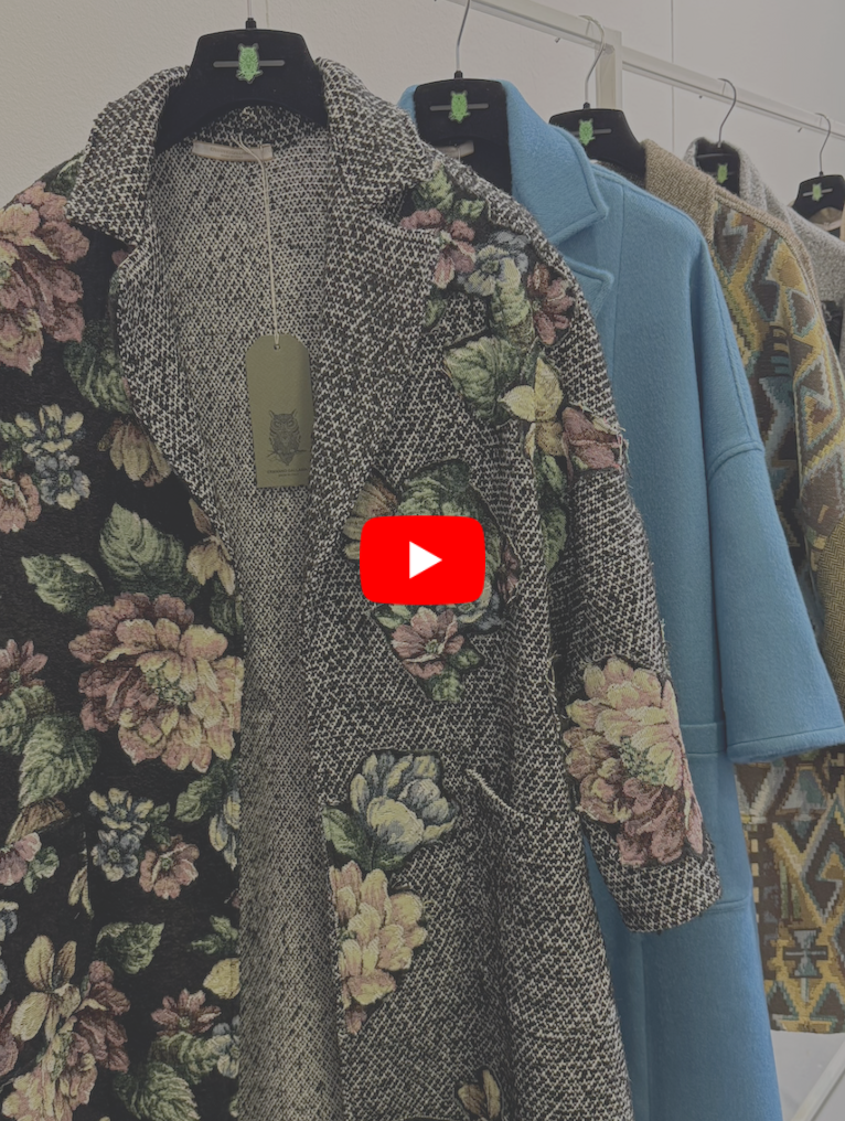 Row of women’s coats hanging on a rack, including a floral tapestry coat in the foreground, with solid blue and patterned outerwear behind it.