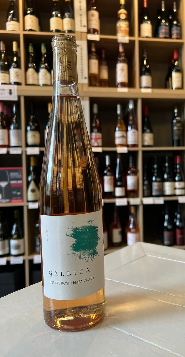 Bottle of Gallica Estate Rosé from Napa Valley displayed on a white counter inside a wine shop, with shelves of assorted wine bottles softly blurred in the background. The pale blush rosé and minimalist label create an elegant, refined presentation.