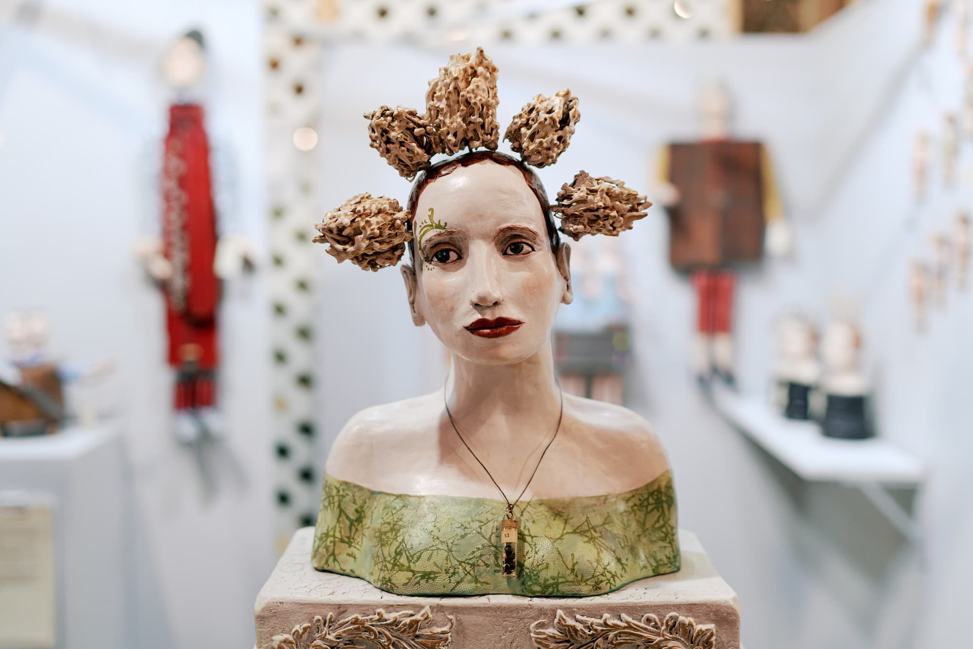 A ceramic bust sculpture of a woman is displayed in an art gallery. The figure has a calm, expressive face and wears an off-the-shoulder green garment, with textured, flower-like forms arranged in her hair and a small pendant necklace, set against softly blurred folk art pieces in the background.