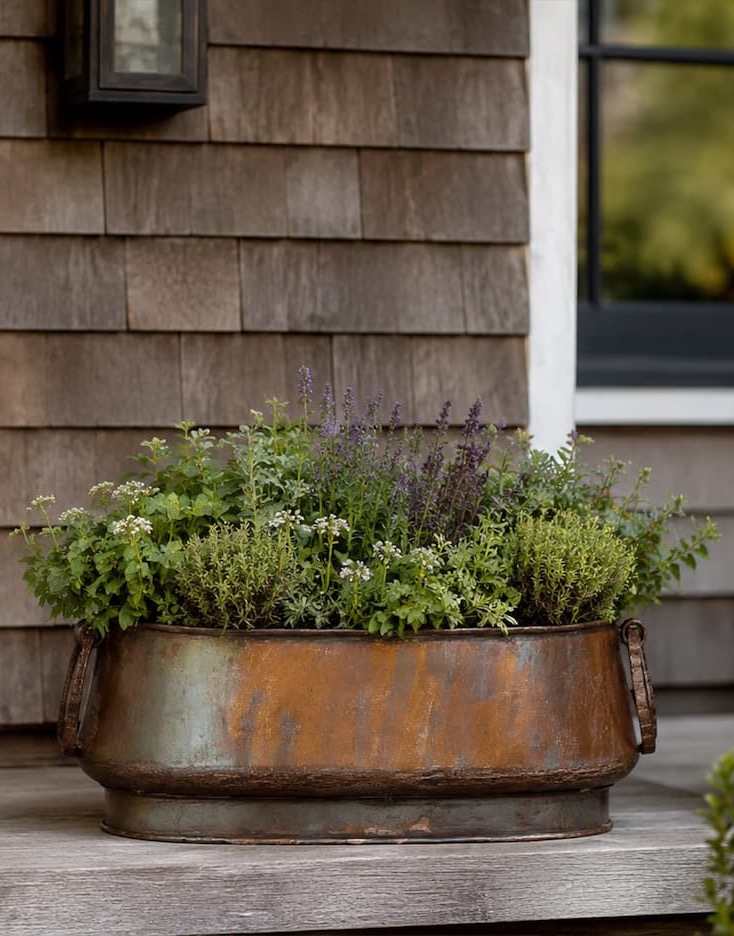 Oval metal planter with a weathered patina, filled with mixed herbs and greenery, placed on a wooden porch against a shingled exterior wall.