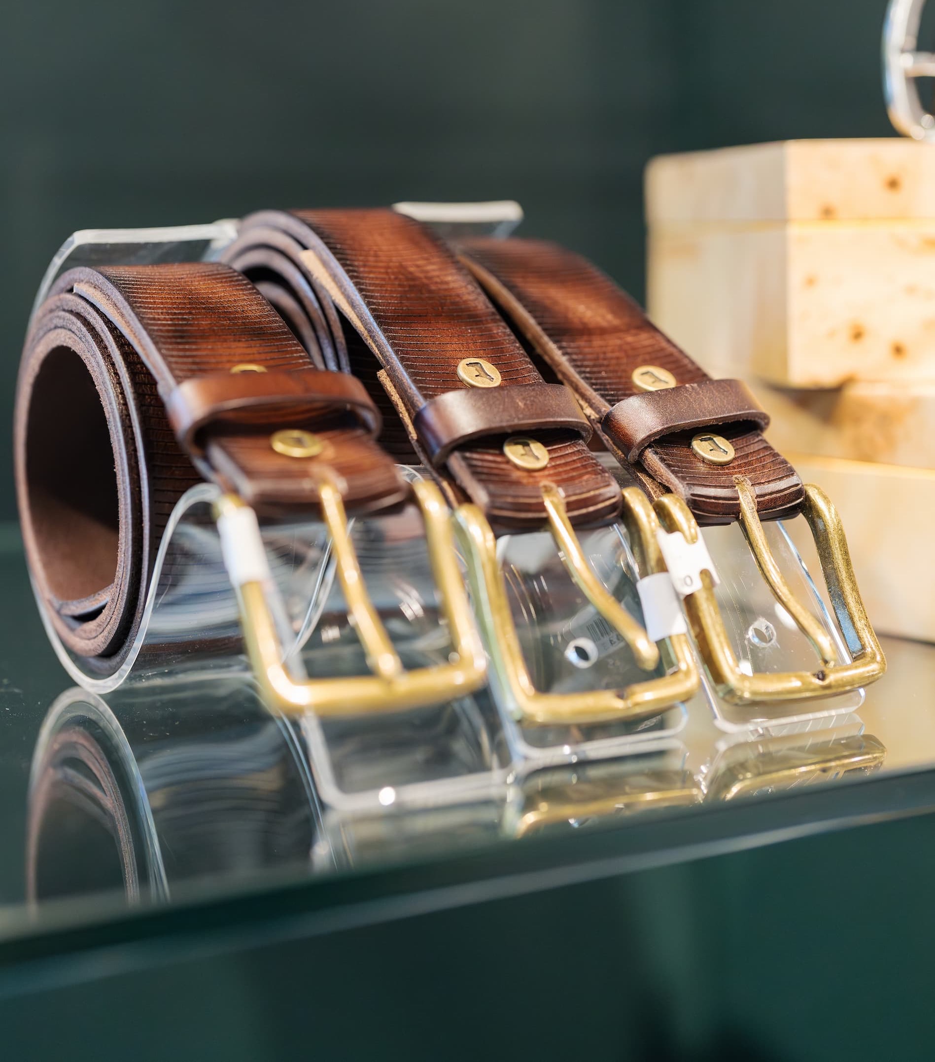 Display of brown leather belts neatly arranged on a glass shelf, each featuring brass buckles and subtle texture. The polished hardware and warm tones create a classic, well-crafted look, with soft reflections visible on the surface.