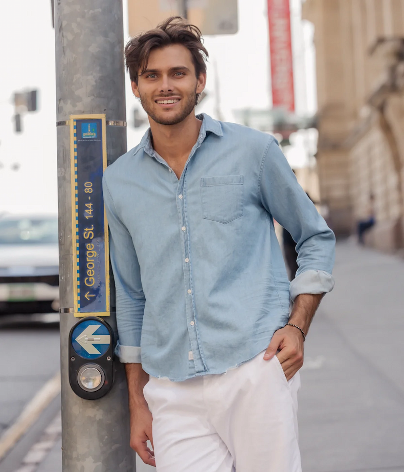 Man wearing a light blue button-down shirt and white pants, standing on a city sidewalk. Fearrington Village.