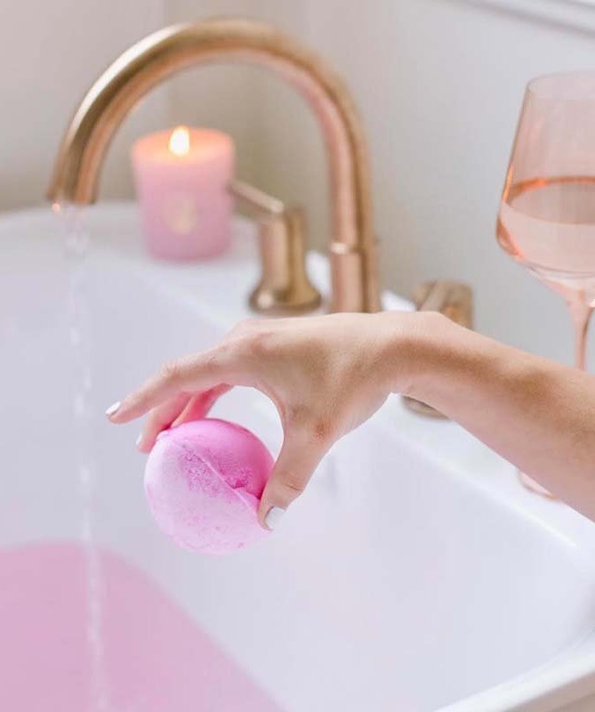 Pink bath bomb being dropped into a bathtub beside a glowing candle and glass of rosé. Fearrington Village.