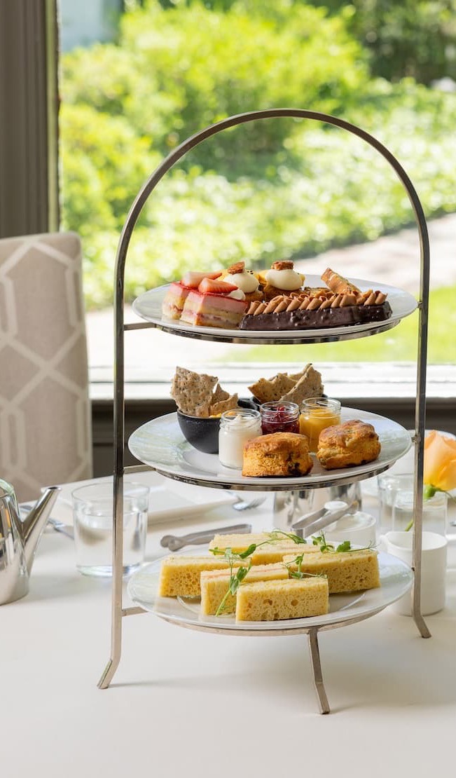 Elegant three-tier afternoon tea stand featuring finger sandwiches, fresh scones with preserves and cream, and an assortment of pastries and desserts, set on a white tablecloth beside a silver teapot and garden-view windows.