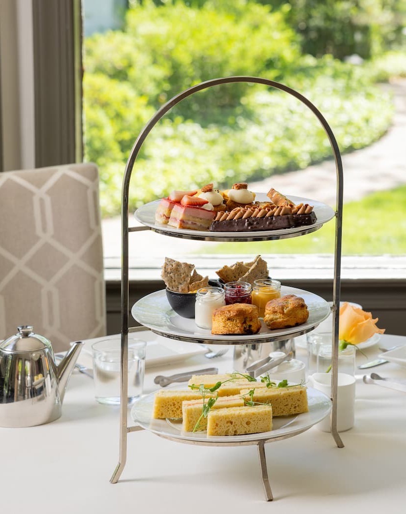 Elegant three-tier afternoon tea stand featuring finger sandwiches, fresh scones with preserves and cream, and an assortment of pastries and desserts, set on a white tablecloth beside a silver teapot and garden-view windows.