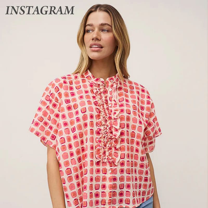 Woman wearing a short-sleeve blouse with a pink and coral geometric print, featuring ruffle detailing down the button-front placket and a stand collar. Styled against a neutral backdrop with blue jeans partially visible.