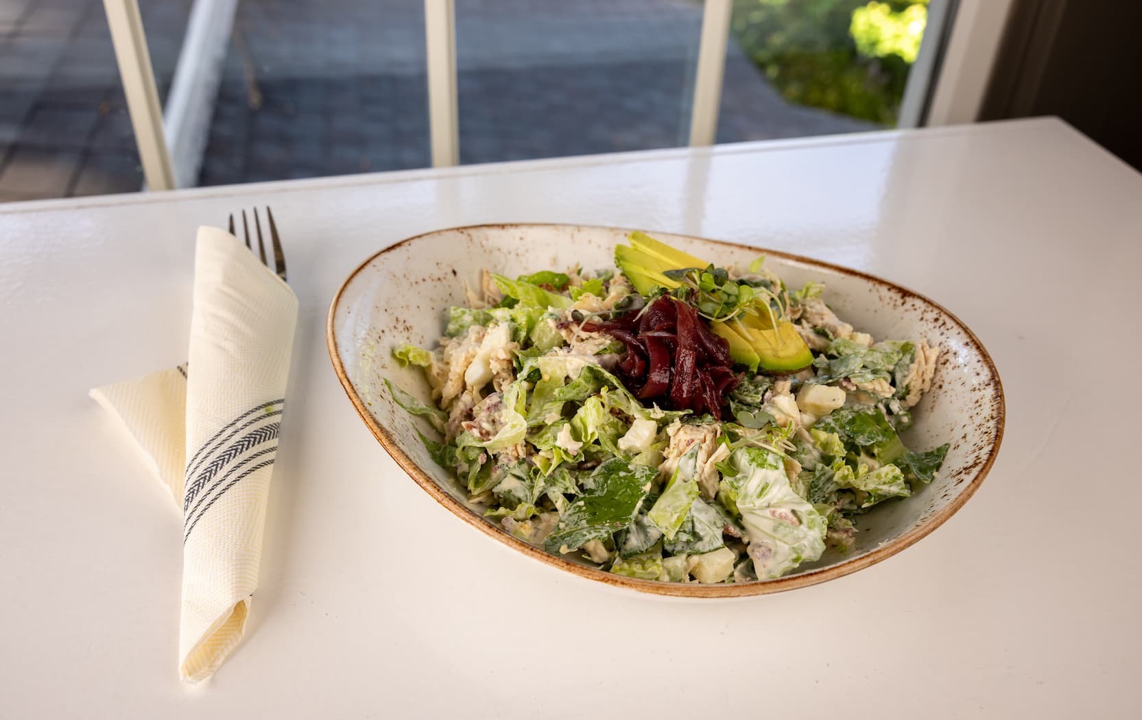 A bowl of creamy chicken salad sits on a white table by a window, topped with avocado slices, pickled onions, and microgreens, with a folded napkin and fork set neatly to the side.