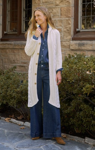 Woman wearing a long cream cable-knit cardigan over a denim shirt and wide-leg jeans, standing outside a stone building. Fearrington Village.