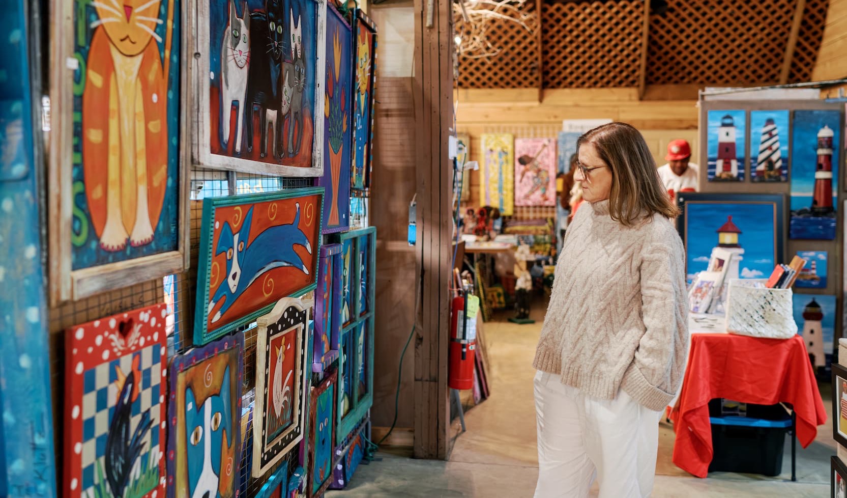 A woman stands inside a rustic art market or gallery, studying a wall of brightly colored folk art paintings featuring animals and playful designs. She wears a light sweater and white pants, with additional artwork and vendor tables visible in the warmly lit space behind her.