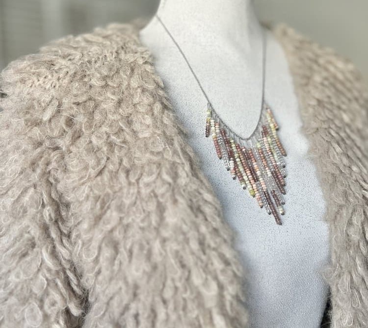 Mannequin dressed in a textured, curly faux-fur jacket, styled with a delicate, multicolored beaded fringe necklace draped across the neckline.