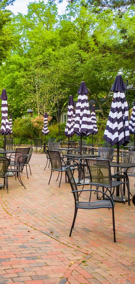 Outdoor patio with brick paving, metal café tables and chairs, and multiple closed black-and-white striped umbrellas set among leafy trees.